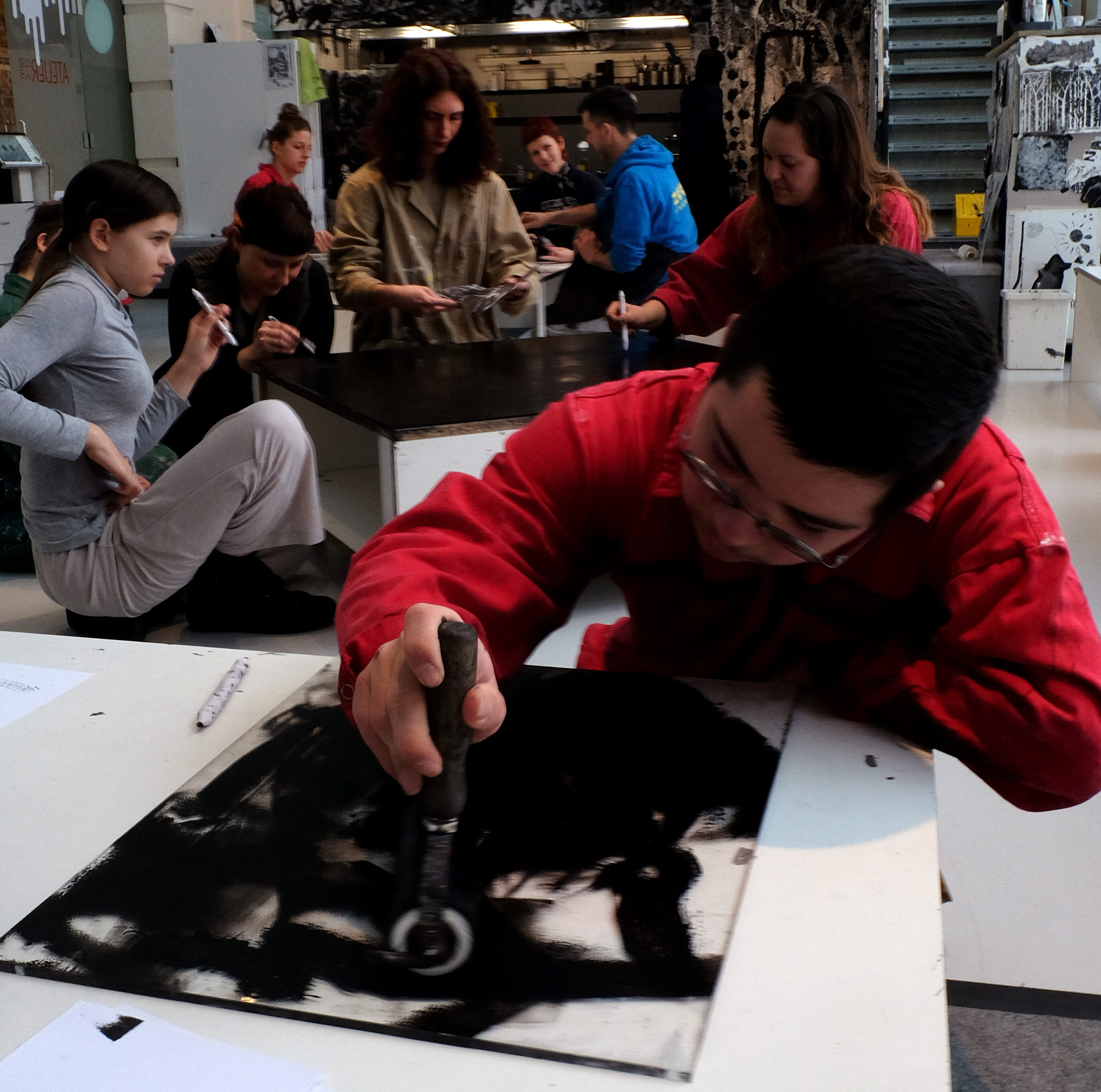 Eine Gruppe junger Menschen arbeitet in einem Kunstatelier. Im Vordergrund beugt sich eine Person in einem roten Overall mit Schutzbrille über einen Tisch und bearbeitet konzentriert eine schwarz bemalte Fläche mit einem Rundwerkzeug. Im Hintergrund sitzen und stehen weitere Teilnehmende an einem langen Tisch und arbeiten an eigenen Projekten. An der Wand hängt ein großes schwarz-weißes Kunstwerk. Regale mit Materialien und Kisten füllen den Hintergrund