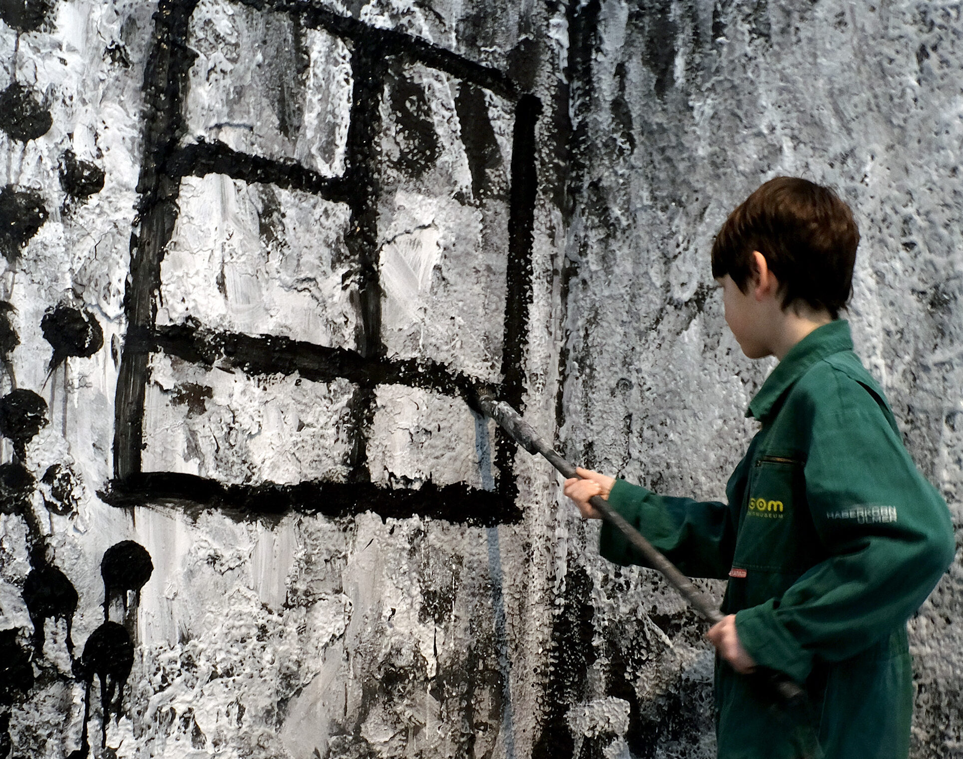 Ein Kind mit kurzem braunem Haar trägt einen grünen Overall und malt mit einem langen Pinsel schwarze Linien auf eine großflächige, schwarz-weiß gestaltete Wand. Die Linien formen ein gitterartiges Muster. Zu den Füßen steht ein schwarzer Eimer mit Farbe. Der Boden ist mit Farbspuren bedeckt. Die Szene wirkt wie eine begehbare Kunstinstallation.