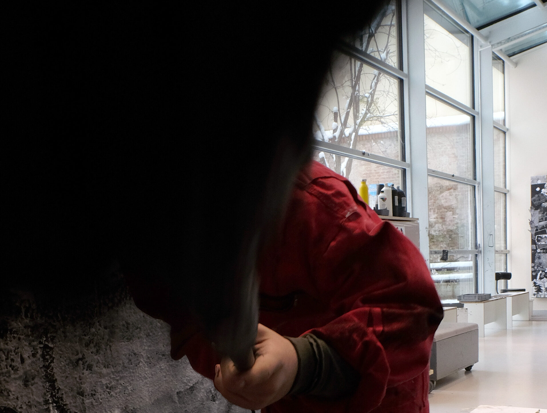 Eine Person in einem roten Overall steht vor einer großen, schwarz-weiß bemalten Wand und arbeitet mit einem Werkzeug oder Pinsel daran. Der Boden ist mit Farbspritzern bedeckt. Im Hintergrund ist ein heller, moderner Atelierraum mit großen Fensterfronten und weiteren Kunstwerken zu sehen