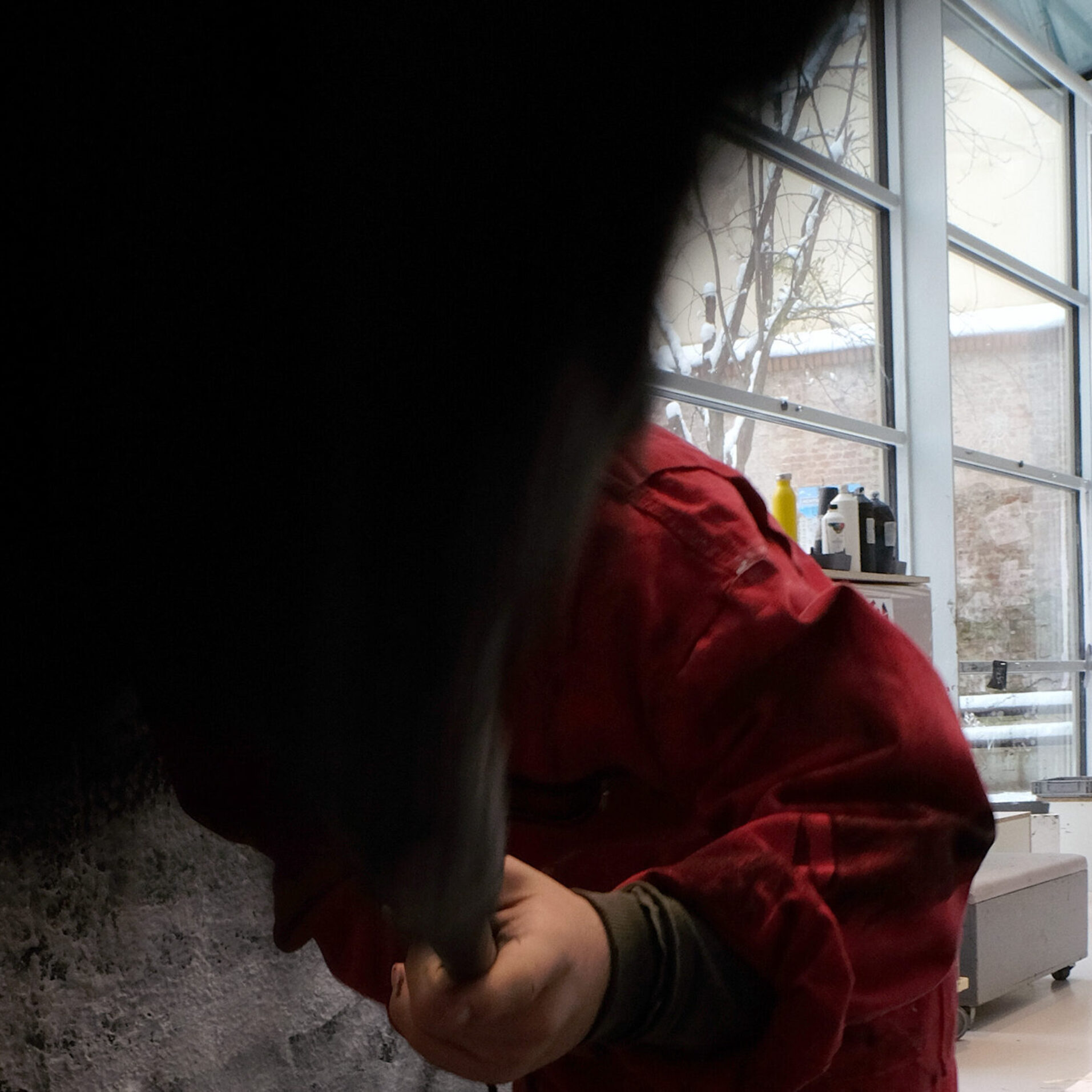 Eine Person in einem roten Overall steht vor einer großen, schwarz-weiß bemalten Wand und arbeitet mit einem Werkzeug oder Pinsel daran. Der Boden ist mit Farbspritzern bedeckt. Im Hintergrund ist ein heller, moderner Atelierraum mit großen Fensterfronten und weiteren Kunstwerken zu sehen