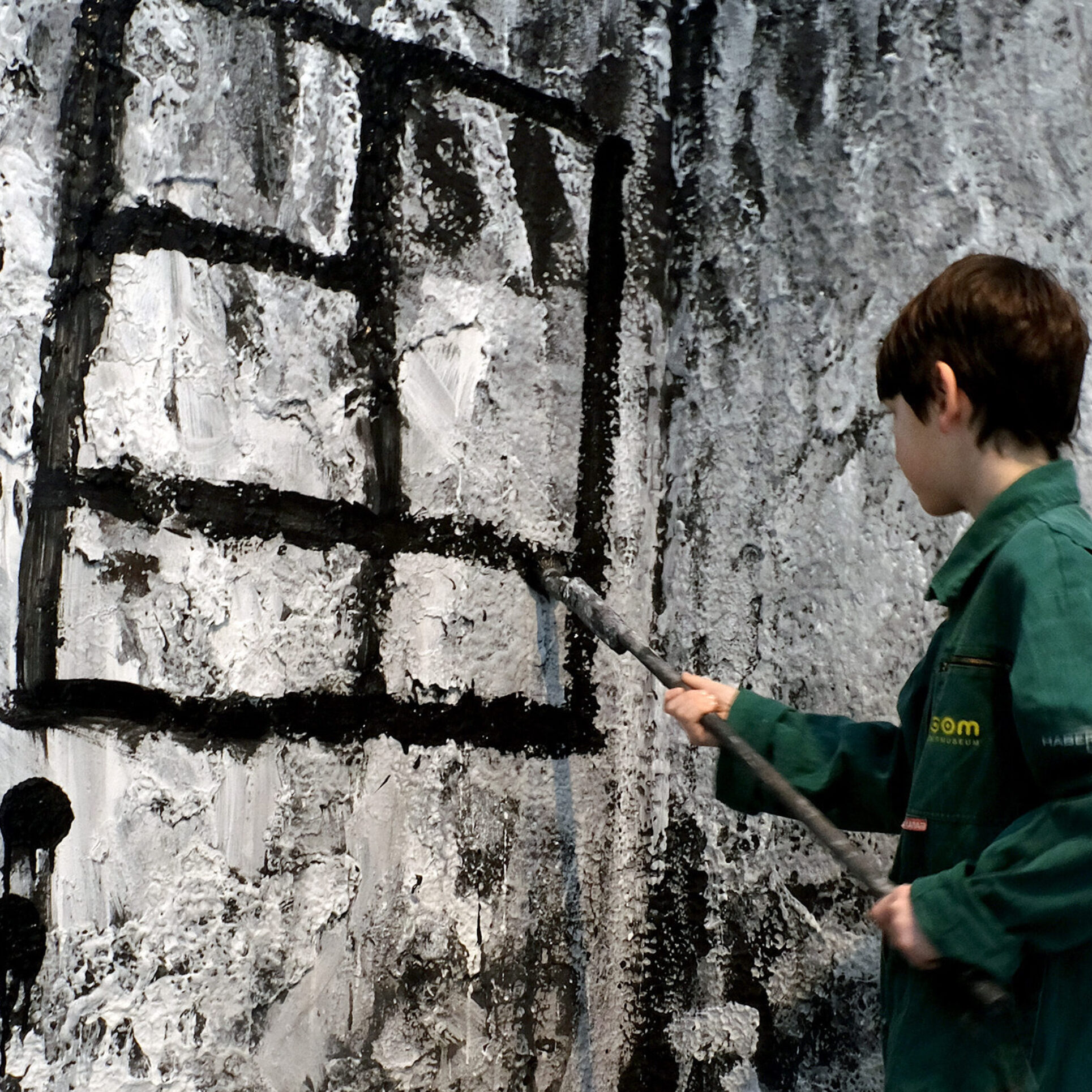 Ein Kind mit kurzem braunem Haar trägt einen grünen Overall und malt mit einem langen Pinsel schwarze Linien auf eine großflächige, schwarz-weiß gestaltete Wand. Die Linien formen ein gitterartiges Muster. Zu den Füßen steht ein schwarzer Eimer mit Farbe. Der Boden ist mit Farbspuren bedeckt. Die Szene wirkt wie eine begehbare Kunstinstallation.