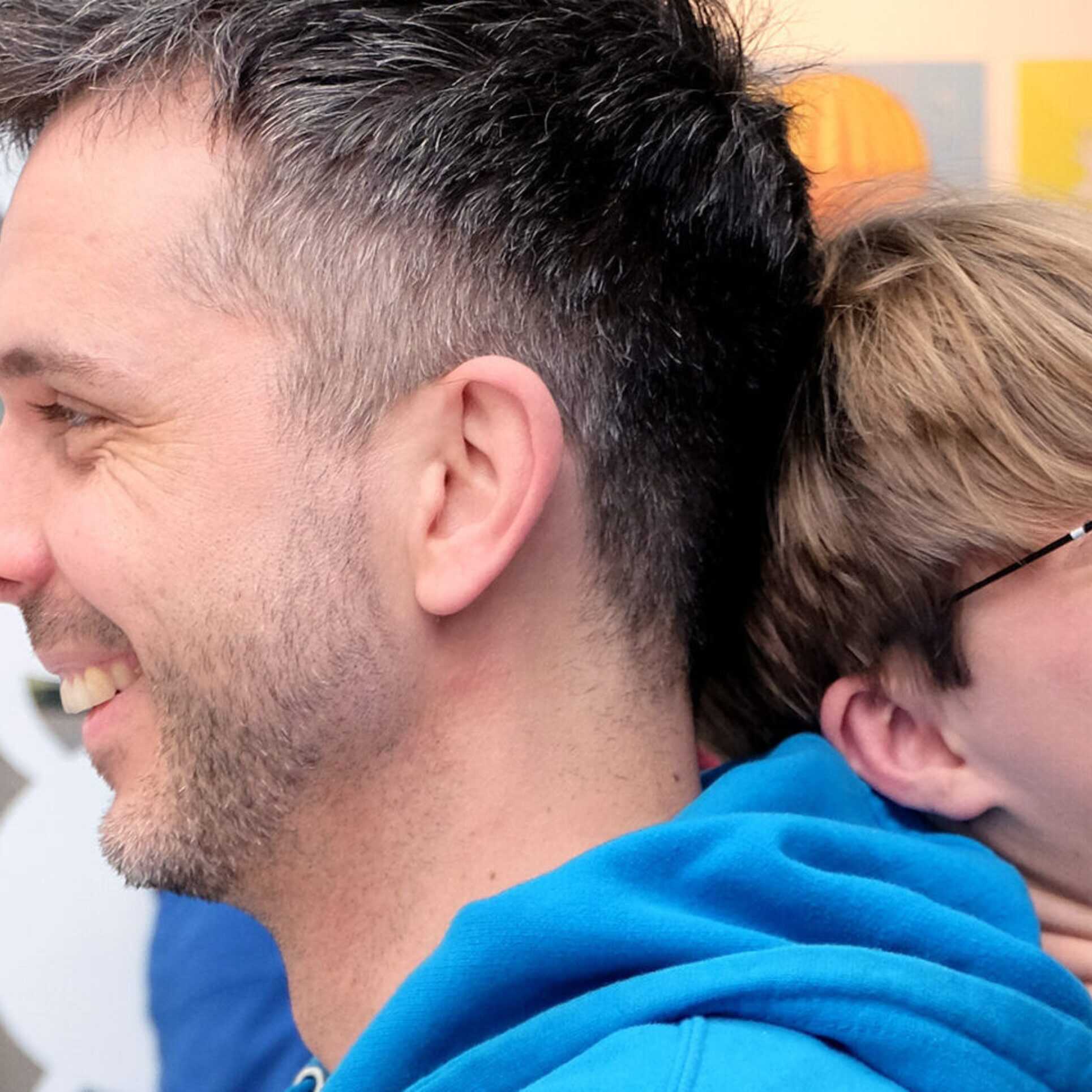 Ein erwachsener Mann mit dunklem Haar und Dreitagebart trägt einen blauen Hoodie. Er steht Rücken an Rücken mit einem Jugendlichen mit blonder Brille und schwarzem Pullover. Beide lachen und wirken fröhlich. Im Hintergrund befindet sich ein moderner, lichtdurchfluteter Raum mit Glaswänden, in dem weitere Personen zu sehen sind.