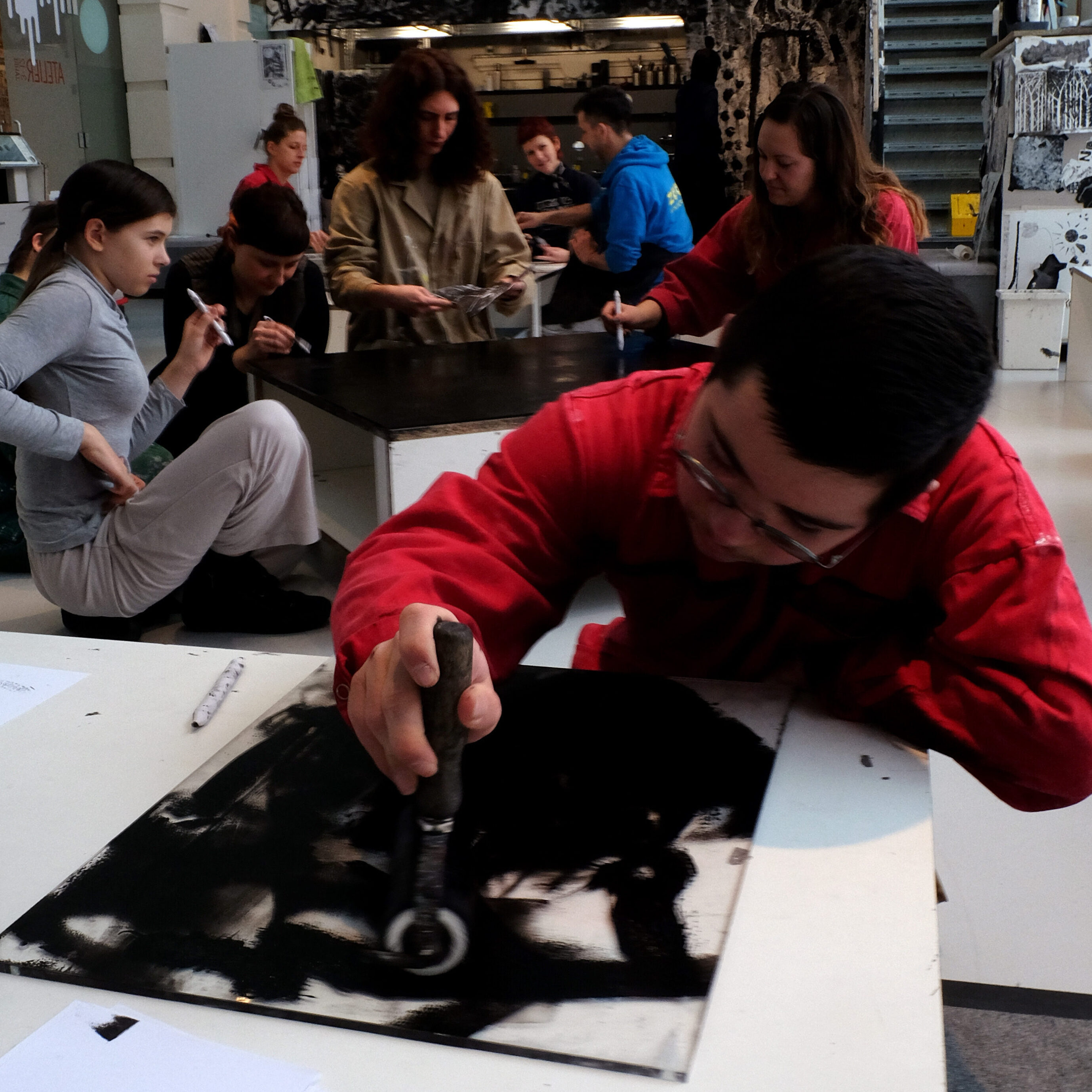 Eine Gruppe junger Menschen arbeitet in einem Kunstatelier. Im Vordergrund beugt sich eine Person in einem roten Overall mit Schutzbrille über einen Tisch und bearbeitet konzentriert eine schwarz bemalte Fläche mit einem Rundwerkzeug. Im Hintergrund sitzen und stehen weitere Teilnehmende an einem langen Tisch und arbeiten an eigenen Projekten. An der Wand hängt ein großes schwarz-weißes Kunstwerk. Regale mit Materialien und Kisten füllen den Hintergrund
